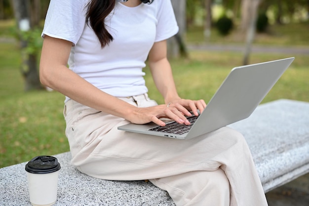 Scatto ritagliato Giovane donna asiatica che usa il suo laptop portatile sulla panchina nel parco cittadino