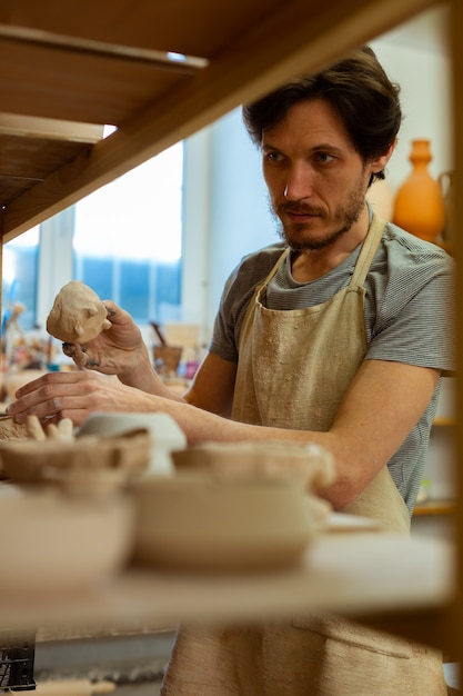 Scelta di pentole adatte. Maestro professionista dell'argilla dai capelli scuri che sistema la sua collezione di pentole rimanendo vicino agli scaffali