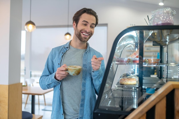 Scelta. Giovane uomo barbuto con un sorriso a trentadue denti che tiene il caffè che punta con la mano al banco di mostra del cibo nella caffetteria durante il giorno