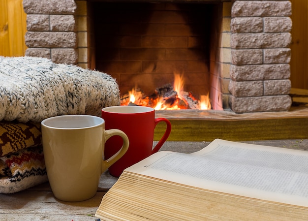 Scena accogliente davanti al camino con due grandi tazze con tè, libro aperto e sciarpa di lana.