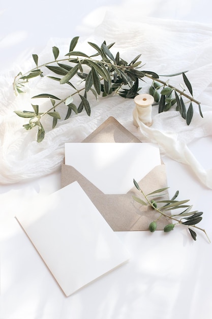 Scena di mockup di cancelleria per matrimoni estivi Busta vuota per biglietti di auguri con rami di ulivo e nastro di seta Sfondo bianco con runner da tavolo in cotone nelle ombre del sole Vista dall'alto piatta verticale