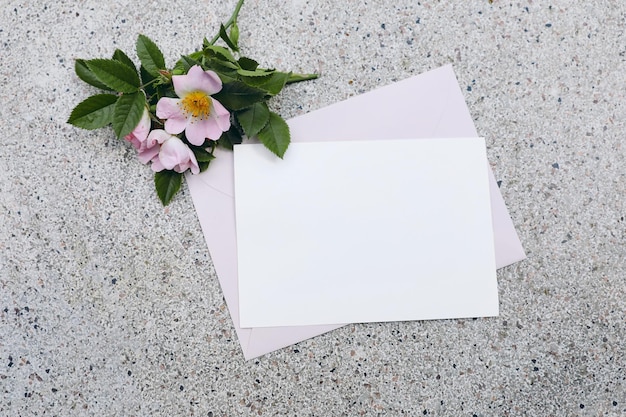 Scena di mockup di cancelleria per matrimoni estivi Invito biglietto di auguri vuoto orizzontale busta rosa e fiori di rosa canina in fiore su sfondo terrazzo strutturato Vista dall'alto piatta Nessuna gente