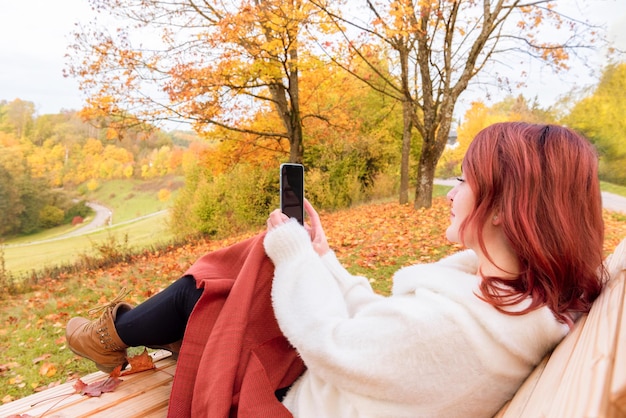 Scenario autunnale con una giovane donna seduta su una panchina che usa il suo smartphone coperto da una coperta