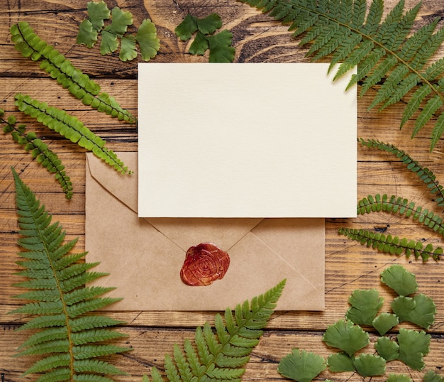 Scheda di carta bianca con busta sigillata e sigillo di cera rossa posata su un tavolo di legno marrone con foglie di felce intorno alla vista dall'alto. Scena di mock-up tropicale con carta di invito piatta