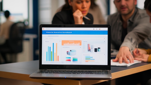 Schermo del laptop pronto per la presentazione del progetto finanziario posizionato sulla scrivania mentre gli uomini d'affari lavorano in background. Diversi dipendenti riuniti in co-working, processo di lavoro in un'azienda impegnata, aiuto nel lavoro di squadra
