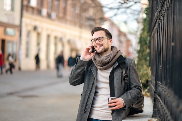 Sciarpa da portare allegra del giovane che comunica sul telefono e che beve caffè sulla via.