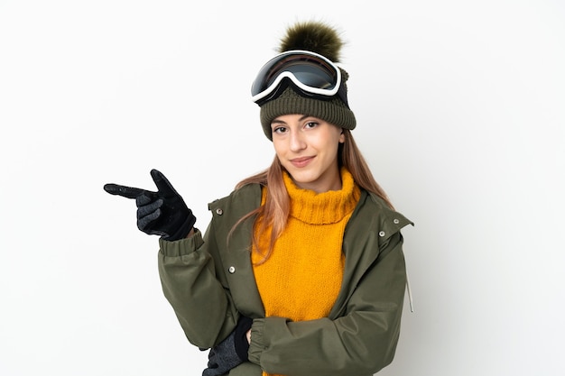 Sciatore donna caucasica con occhiali da snowboard isolati su sfondo bianco che punta il dito a lato