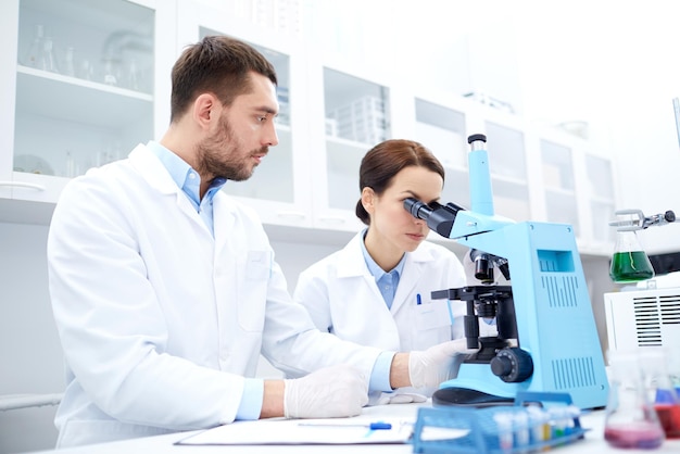 scienza, chimica, tecnologia, biologia e concetto di persone - giovani scienziati con microscopio che fanno test o ricerca in laboratorio clinico e prendono appunti