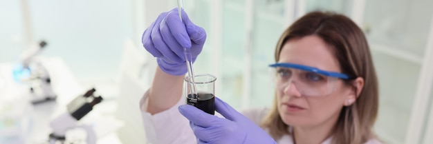 Scienziata focalizzata in occhiali mescola liquido nero in vetreria donna in guanti di gomma lo fa