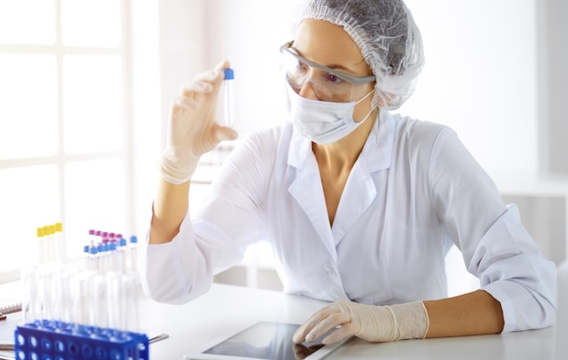 Scienziata professionista in occhiali protettivi che ricerca tubo con reagenti in laboratorio soleggiato. Medicina e ricerca.