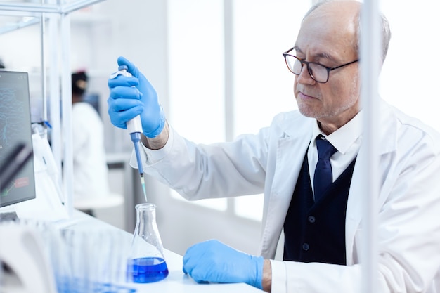 Scienziato anziano che fa un esperimento genetico in un laboratorio affollato usando un contagocce sterile con liquido blu. Chimico professionista senior che utilizza pipetta con soluzione blu per test microbiologici.