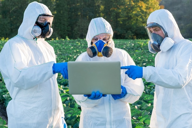 Scienziato e agronomi che indossano un equipaggiamento protettivo bianco, una maschera chimica e occhiali usano il laptop sul campo dell'azienda agricola.