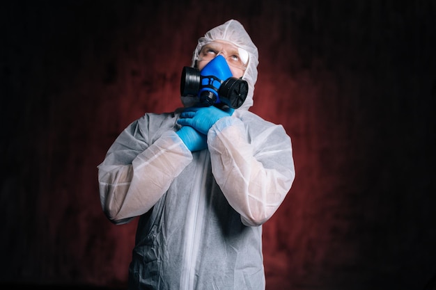 Scienziato epidemiologo in hazmat protettivo, occhiali e respiratore si sta strangolando con le mani. Concetto di pandemia di Coronavirus COVID-19. Studio girato su sfondo rosso scuro isolato.