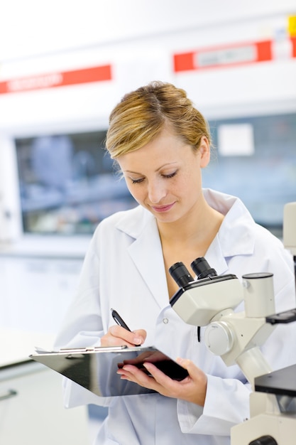 Scienziato femminile serio che scrive sulla sua lavagna per appunti davanti ad un microscopio