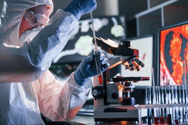 Scienziato in uniforme protettiva bianca vicino a display con dati funziona con coronavirus e provette ematiche in laboratorio