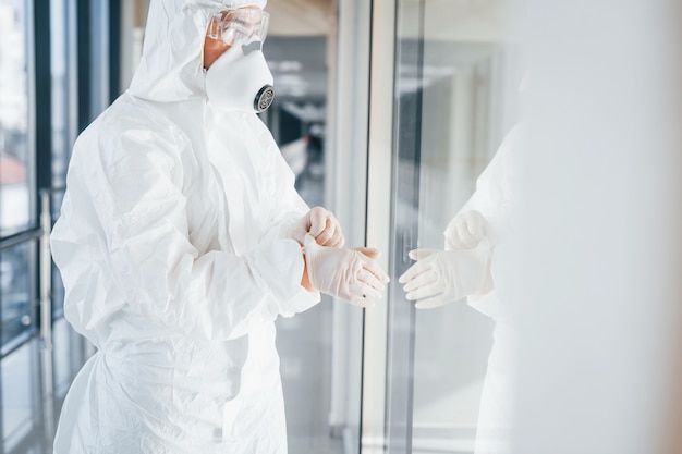 Scienziato medico femminile in camice da laboratorio, occhiali difensivi e maschera in piedi all'interno e indossando guanti