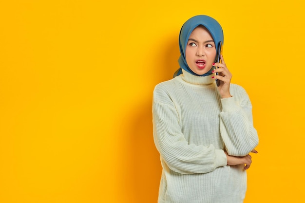 Scioccata bella donna asiatica in maglione bianco e hijab che parla al cellulare mentre guarda da parte isolata su sfondo giallo