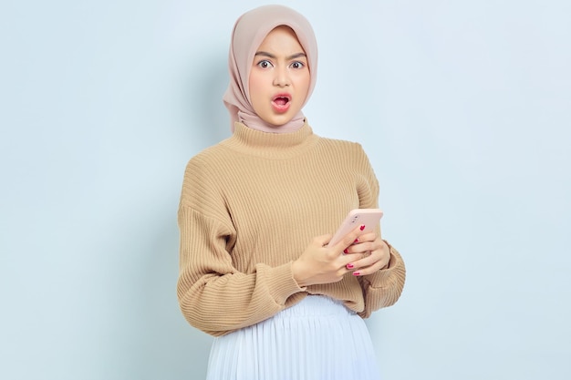 Scioccata bella donna musulmana asiatica in maglione marrone utilizzando il telefono cellulare ha ricevuto buone notizie isolate su sfondo bianco Concetto di stile di vita religioso della gente