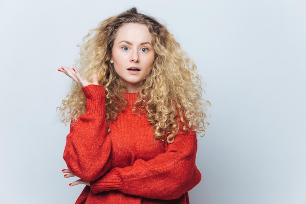 Scioccata bella giovane donna bionda dagli occhi azzurri guarda con stupore alla telecamera indossa un maglione rosso sciolto essendo insoddisfatto di qualcosa di isolato su sfondo bianco Concetto di stupore