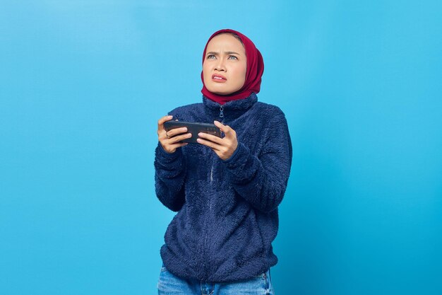 Scioccata giovane donna asiatica che gioca a un videogioco su uno smartphone e guarda in alto su sfondo blu