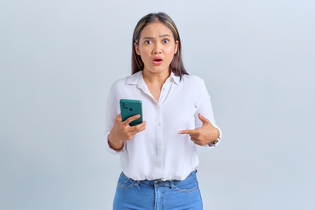 Scioccata giovane donna asiatica che punta le dita al telefono cellulare e guardando la fotocamera isolata su sfondo bianco