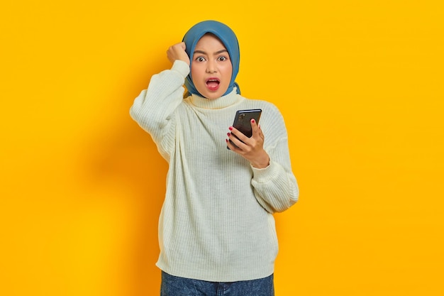 Scioccata giovane donna asiatica in maglione bianco e hijab utilizzando sul telefono cellulare e tenendo la testa con la mano isolata su sfondo giallo