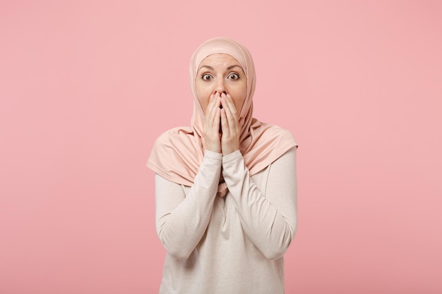 Scioccata giovane donna musulmana araba in abiti leggeri hijab in posa isolata su sfondo rosa ritratto in studio. Concetto di stile di vita dell'Islam religioso della gente. Mock up spazio di copia. Coprire la bocca con le mani.