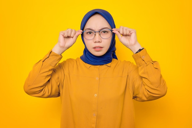 Scioccata giovane donna musulmana asiatica vestita con una camicia arancione che guarda la fotocamera con la mano che tocca i suoi occhiali interessata al grande promo isolato su sfondo giallo