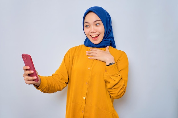 Scioccata giovane donna musulmana asiatica vestita con una camicia arancione che guarda lo schermo del telefono cellulare con la bocca aperta che reagisce a grandi notizie isolate su sfondo bianco