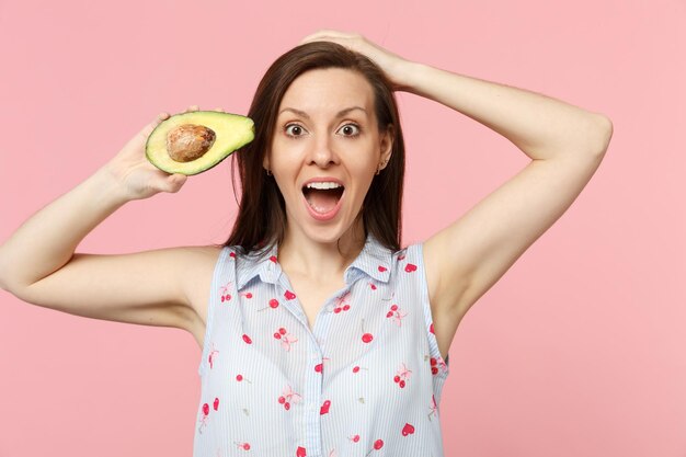 Scioccata giovane ragazza in abiti estivi tenendo la bocca aperta tenere fresco frutto di avocado maturo isolato su sfondo rosa pastello in studio. Lo stile di vita vivido delle persone si rilassa il concetto di vacanza. Mock up spazio di copia.