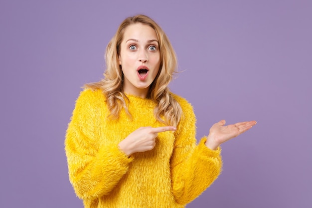 Scioccata ragazza giovane donna in maglione di pelliccia gialla in posa isolata su sfondo viola pastello ritratto in studio. Concetto di stile di vita delle emozioni delle persone. Mock up spazio di copia. Puntando il dito indice da parte.