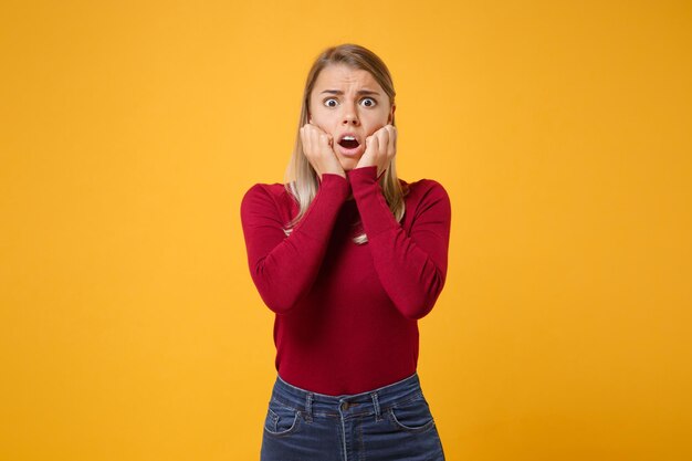 Scioccato disorientato giovane donna bionda ragazza in abiti casual in posa isolato su sfondo giallo arancione ritratto in studio. Concetto di stile di vita di emozioni della gente. Simula lo spazio della copia. Metti le mani sulle guance.