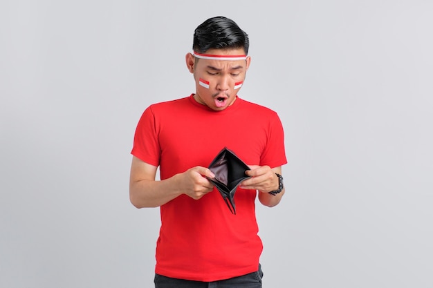 Scioccato giovane asiatico che celebra il giorno dell'indipendenza indonesiana mentre esamina il portafoglio vuoto isolato su sfondo bianco