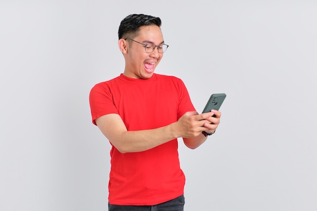 Scioccato giovane asiatico guardando lo schermo del telefono cellulare che reagisce alle notizie online con la bocca aperta isolata su sfondo bianco