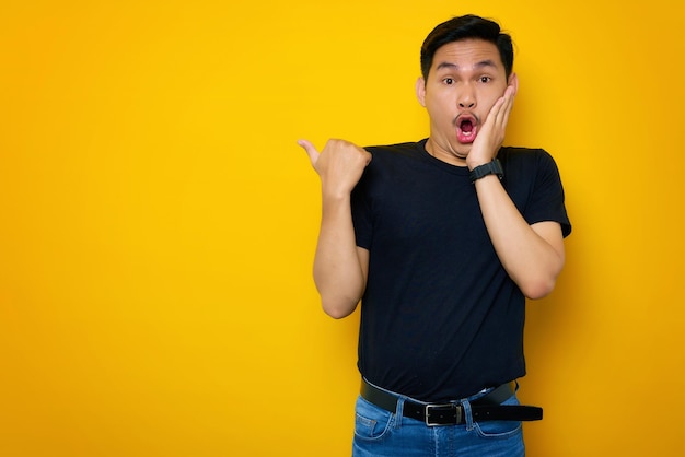 Scioccato giovane asiatico in maglietta casual che punta il pollice da parte allo spazio di copia isolato su sfondo giallo Grande concetto di offerta