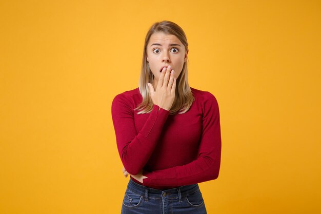 Scioccato giovane donna bionda ragazza in abiti casual in posa isolato su sfondo giallo arancione ritratto in studio. Concetto di stile di vita di emozioni sincere della gente. Simula lo spazio della copia. Coprendo la bocca con la mano.