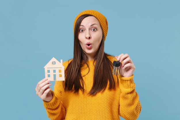 Scioccato giovane donna bruna ragazza in maglione giallo e cappello in posa isolato su sfondo blu ritratto in studio. Persone sincere emozioni concetto di stile di vita. Mock up spazio di copia. Tieni il mazzo di chiavi della casa.