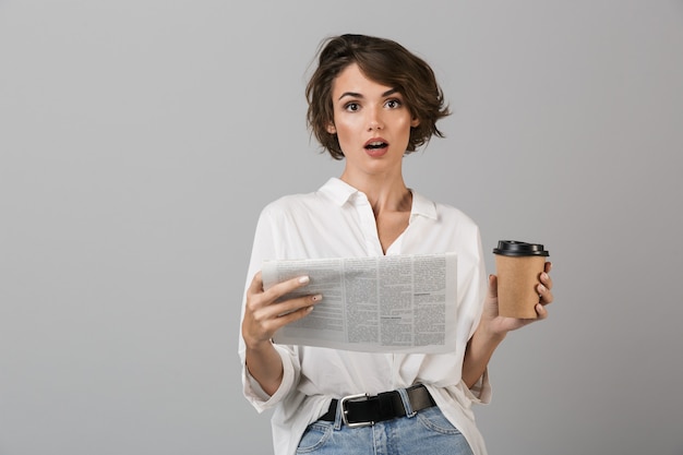 Scioccato giovane donna d'affari in posa isolato sopra il muro grigio leggendo il giornale e bere caffè