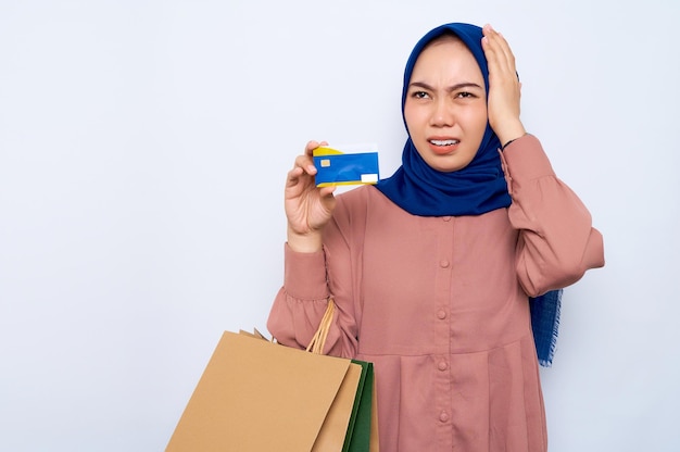 Scioccato giovane donna musulmana asiatica in camicia rosa che tiene borse per pacchi con acquisti dopo lo shopping e carta di credito isolata su sfondo bianco Concetto di stile di vita della gente