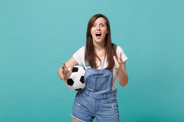 Scioccato giovane donna tifoso di calcio supporta la squadra preferita con pallone da calcio diffondendo le mani isolate su sfondo blu muro turchese. Emozioni della gente, concetto di svago della famiglia di sport. Mock up spazio di copia.