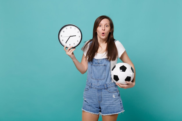 Scioccato giovane tifoso di calcio in possesso di pallone da calcio, orologio rotondo isolato su sfondo blu turchese parete in studio. Il tempo sta finendo. Emozioni della gente, concetto di stile di vita di svago della famiglia di sport.