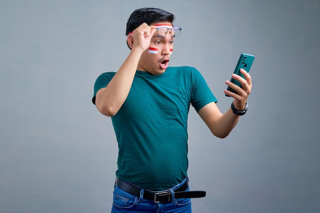 Scioccato giovane uomo asiatico in maglietta casual guardando lo schermo del telefono cellulare togliendosi gli occhiali con la bocca aperta isolati su sfondo grigio concetto di celebrazione del giorno dell'indipendenza indonesiana