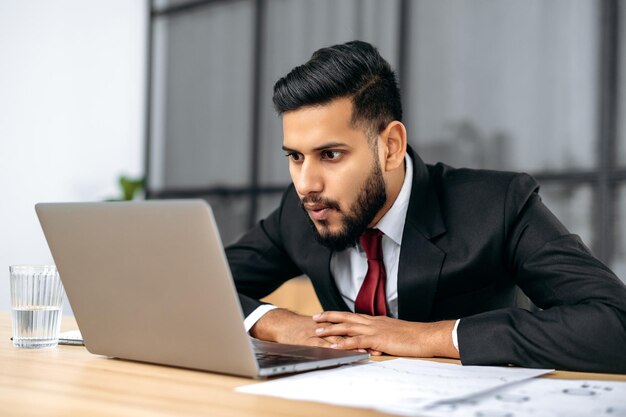 Scioccato perplesso stordito indiano o arabo giovane amministratore delegato della società d'affari in un vestito si siede in un ufficio moderno sguardo sorpreso allo schermo del laptop vedere messaggio inaspettato o notizie sorpreso espressione facciale