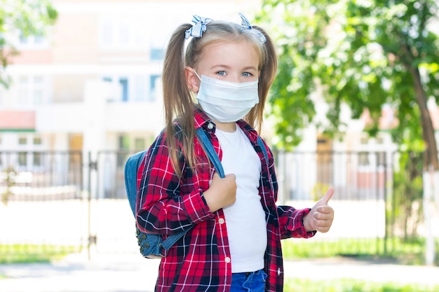 Scolara felice in una maschera protettiva con uno zaino. con una maglietta bianca e una camicia a quadri