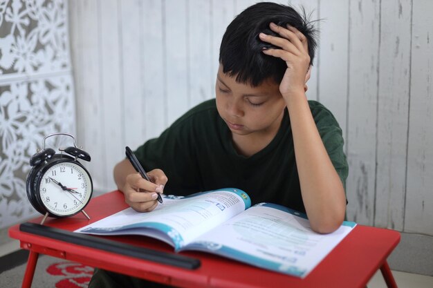 Scolaretto esausto stanco di studiare che tiene la testa con le mani seduto alla scrivania con la carta