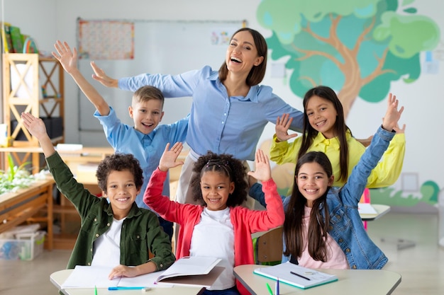 Scolari diversi eccitati che alzano le braccia e sorridono seduti in classe con la loro femmina felice