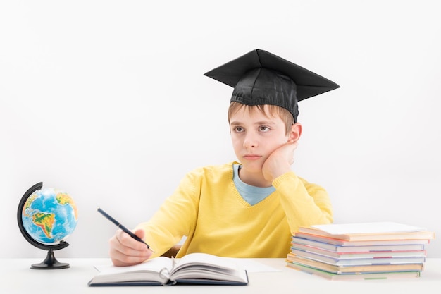 Scolaro elementare annoiato alla lezione Ragazzo nel cappello degli studenti scrive nel taccuino Il bambino è stanco di imparare