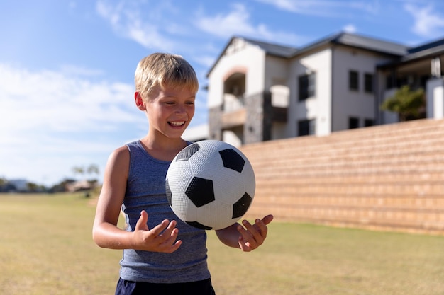 Scolaro elementare caucasico sorridente che gioca con il pallone da calcio sul campo contro l'edificio scolastico