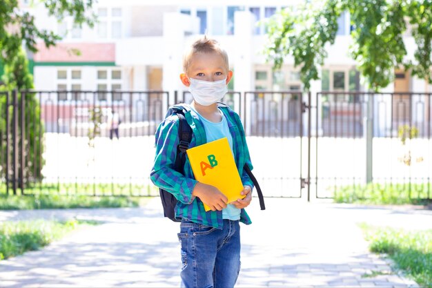 Scolaro in maschera protettiva con uno zaino e un libro di testo in mano. in una maglietta e una camicia a quadri