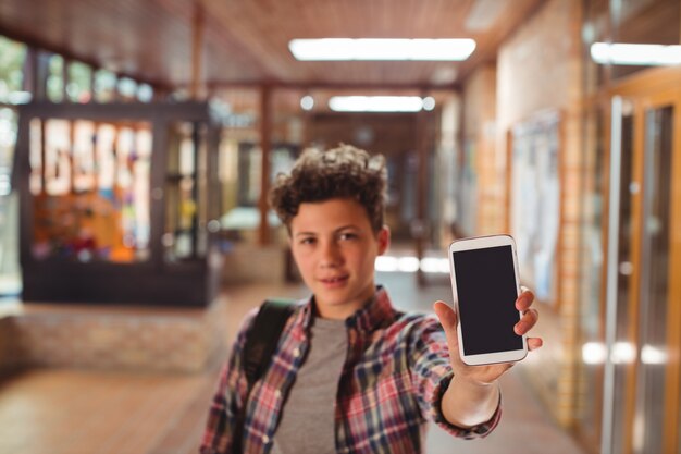 Scolaro in piedi con lo zainetto che mostra il telefono cellulare in corridoio a scuola